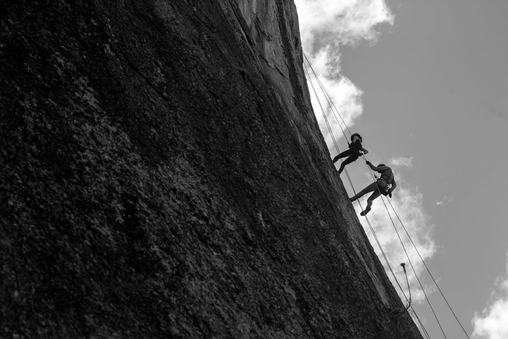 A dramatic black and white shot of two climbers scaling a sheer rock face against a cloudy sky.